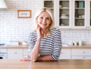 Cheerful middle aged woman standing near table at home kitchen, smile and looking at camera. Portrait of confident joyful female entrepreneur in casual clothes